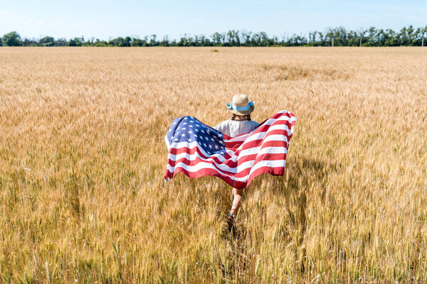 back view of kid in straw hat holding american flag with stars and stripes in field with rye 