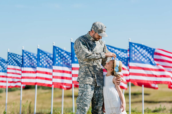 happy military man looking at cute kid in straw hat near american flags with stars and stripes 
