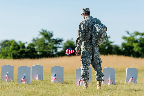back view of military man holding american flag near headstones in graveyard 
