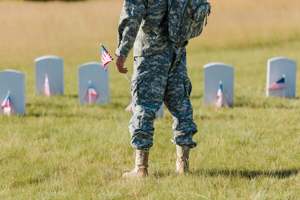 cropped view of military man holding american flag near headstones in graveyard 