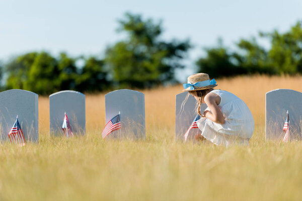 selective focus of child in dress and straw hat  sitting near headstone with american flag in graveyard 