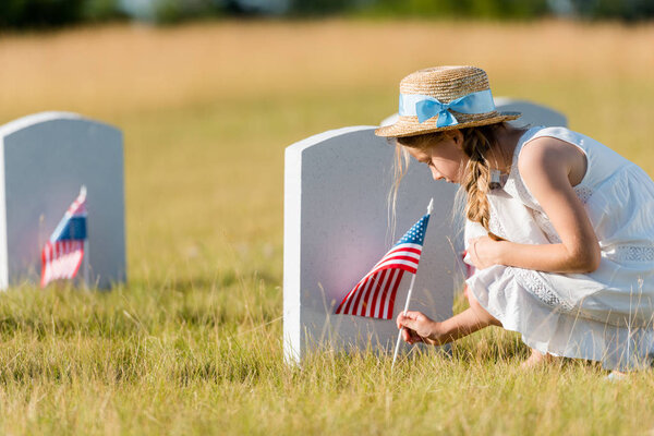 selective focus of adorable kid in straw hat sitting near headstone with american flag in graveyard 