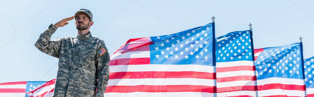 panoramic shot of patriotic soldier in military uniform giving salute near american flags with stars and stripes 