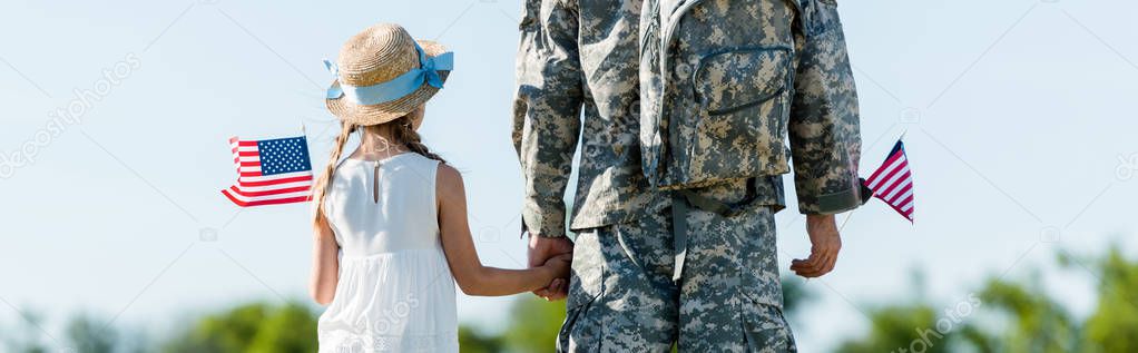 panoramic shot of patriotic child and man in military uniform holding hands and american flags 