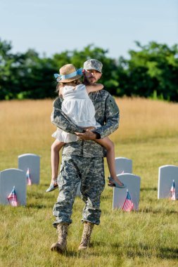 veteran father in cap holding in ams kid in straw hat near headstones with american flags  clipart