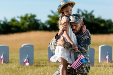 military father in uniform hugging child near headstones in graveyard  clipart