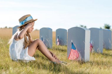 kid in straw hat giving salute while sitting near headstones with american flags  clipart