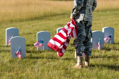 mezarlıkta dururken Amerikan bayrağı tutan askeri üniformalı adamın kırpılmış görünümü 