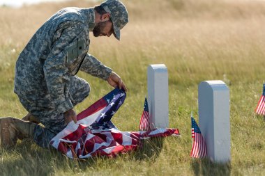 mezarlıkta otururken amerikan bayrağı tutan üniforma ve şapkalı asker 