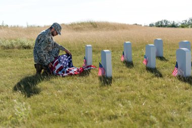 selective focus of soldier in uniform and cap holding american flag while sitting in graveyard  clipart