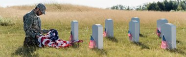 mezarlıkta otururken kamuflaj üniforması ve şapkası amerikan bayrağı tutan üzgün askerin panoramik görüntüsü 