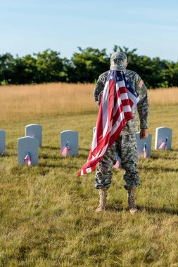  veteran in camouflage uniform covering face with american flag and standing in graveyard  clipart