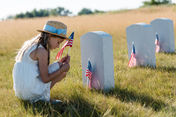 sad kid in straw hat sitting near headstones and holding american flag 