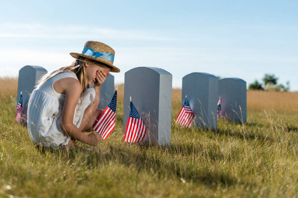 kid in straw hat covering face while sitting near headstones with american flags 