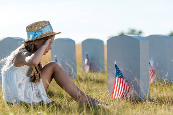 kid in straw hat touching face while sitting near headstones with american flags 