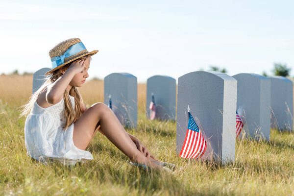 kid in straw hat giving salute while sitting near headstones with american flags 