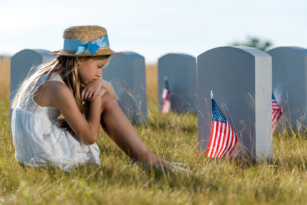 selective focus of sad child sitting and looking at headstones with american flags 