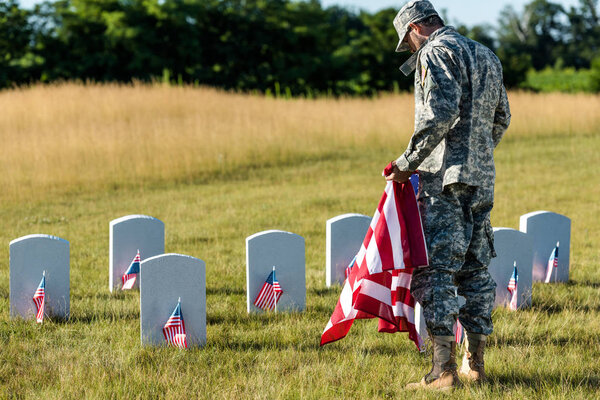military man in uniform and cap holding american flag in graveyard 