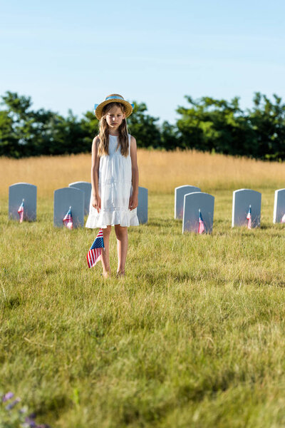 selective focus of child in white dress standing on graveyard with american flag 