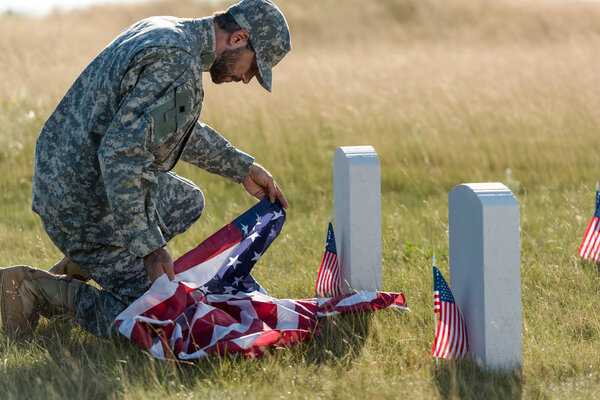 soldier in uniform and cap holding american flag while sitting in graveyard 