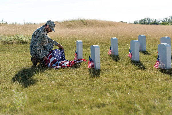 selective focus of soldier in uniform and cap holding american flag while sitting in graveyard 