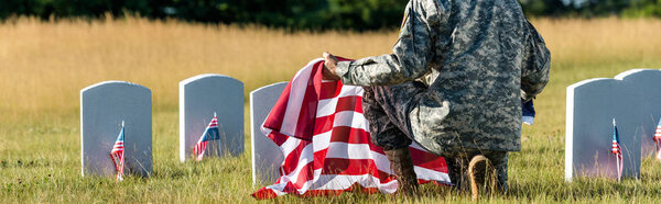 panoramic shot of man in military uniform holding american flag while sitting in graveyard 