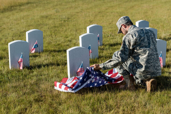 soldier in camouflage uniform and cap holding american flag while sitting in graveyard 