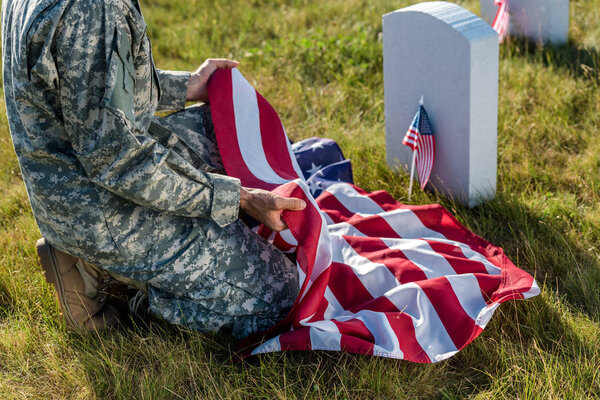 cropped view of soldier in camouflage uniform holding american flag and sitting in graveyard 