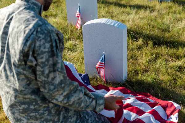selective focus of soldier in camouflage uniform holding american flag and sitting in graveyard 
