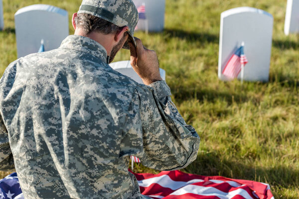 back view of soldier in camouflage uniform touching cap while sitting in graveyard 