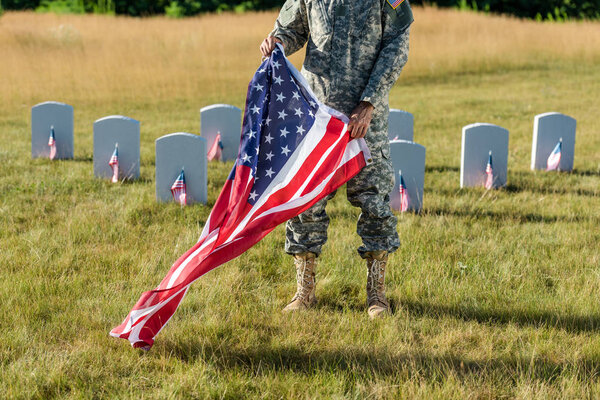 cropped view of man in camouflage uniform holding american flag and standing in graveyard 