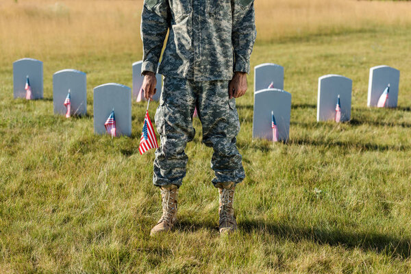 cropped view of veteran in camouflage uniform holding american flag and standing in graveyard 