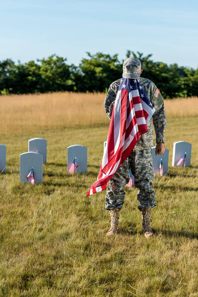  veteran in camouflage uniform covering face with american flag and standing in graveyard 