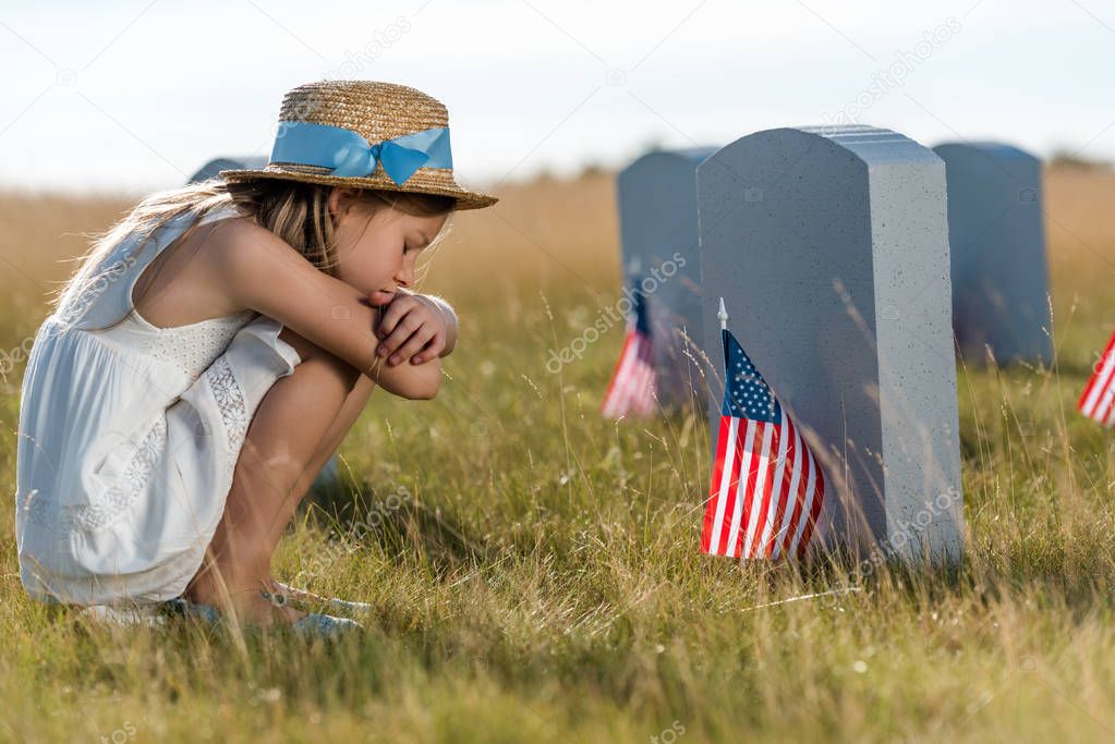sad kid in straw hat sitting near headstones with american flags 