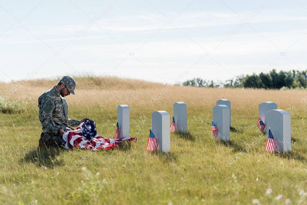 sad soldier in camouflage uniform and cap holding american flag while sitting in graveyard 