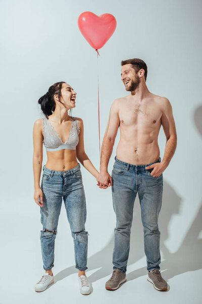 full length view of shirtless couple holding hands and balloon in heart form, and laughing while looking at each other