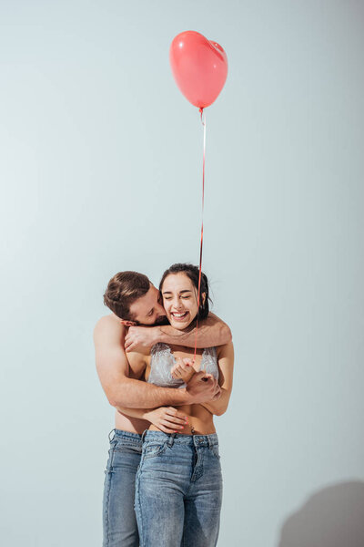muscular man kissing and  hugging girlfriend from back while girl laughing and holding balloon in heart form