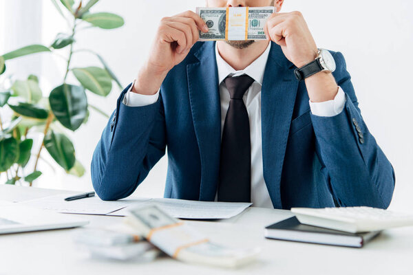 cropped view of man in suit covering face while smelling dollar banknotes