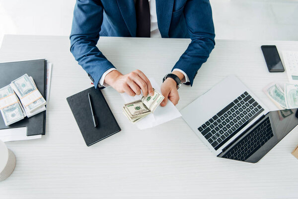 cropped view of businessman holding bribe near notebook and pen on table 