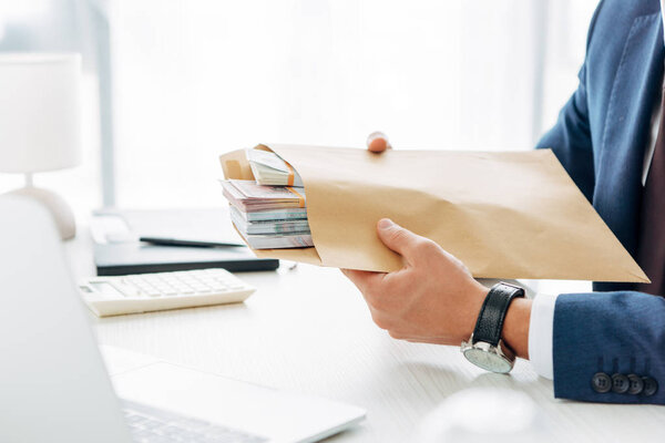 cropped view of businessman holding envelope with money and laptop in office 