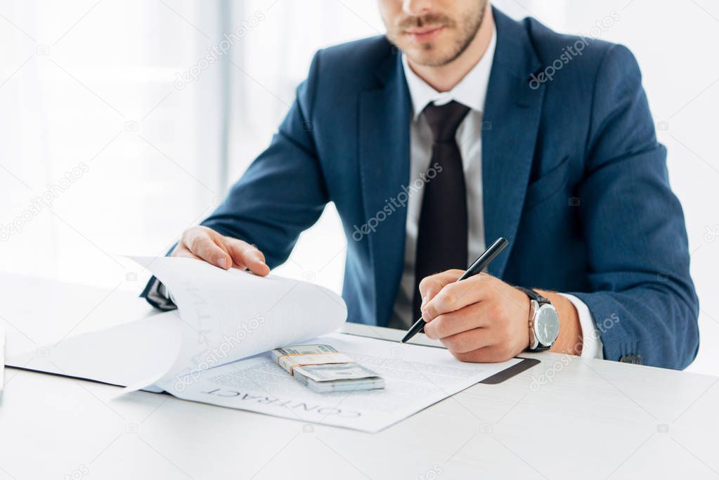 cropped view of man holding pen and contract near bribe on table 