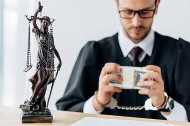 selective focus of statuette of justice near handsome handcuffed judge in glasses holding dollar banknotes  clipart