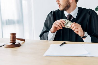 cropped view of judge holding dollar banknotes near gavel, documents and pen  clipart