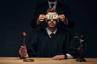 cropped view of man standing and covering face of judge with bribe on black  clipart
