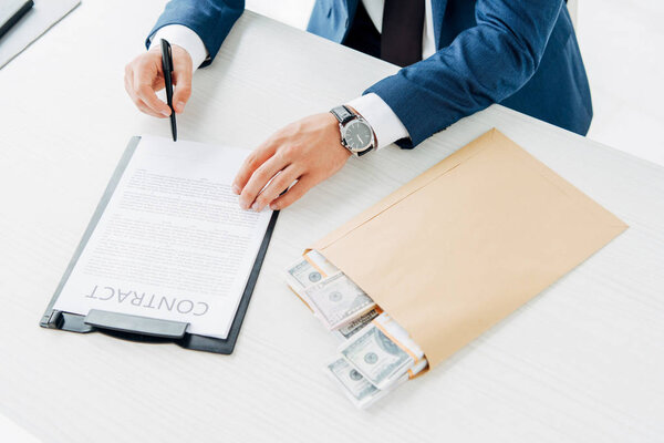 top view of businessman holding pen while signing contract near envelope with money on table 