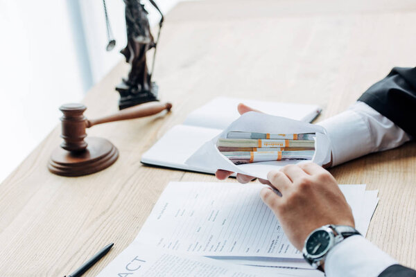 cropped view of judge holding envelope with money near gavel and statuette of justice 