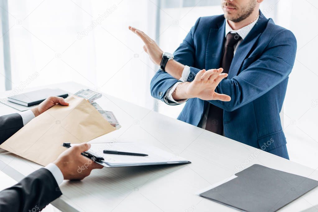 cropped view of businessman gesturing near business partner giving envelope with bribe and clipboard with contract in office 