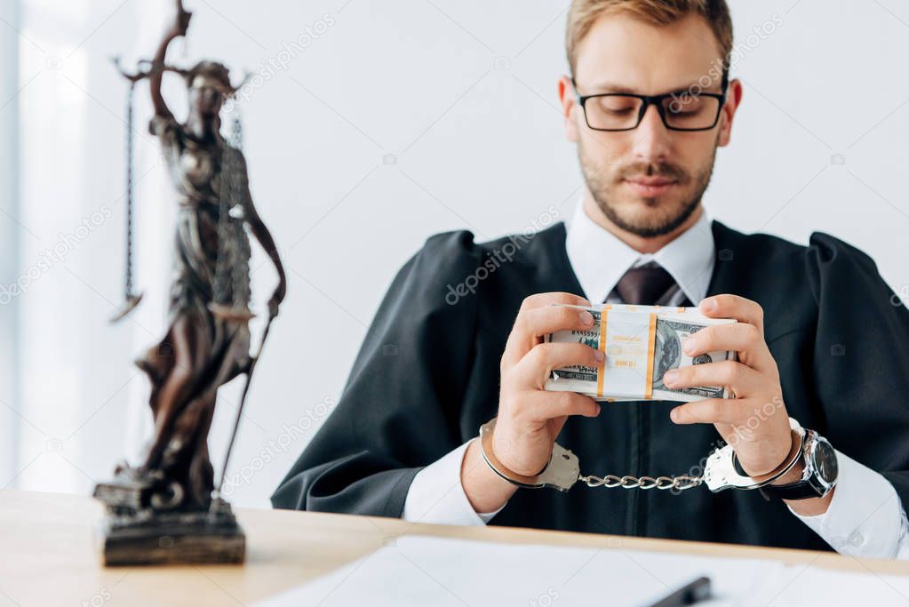 selective focus of handsome handcuffed judge in glasses holding dollar banknotes near statuette of justice 