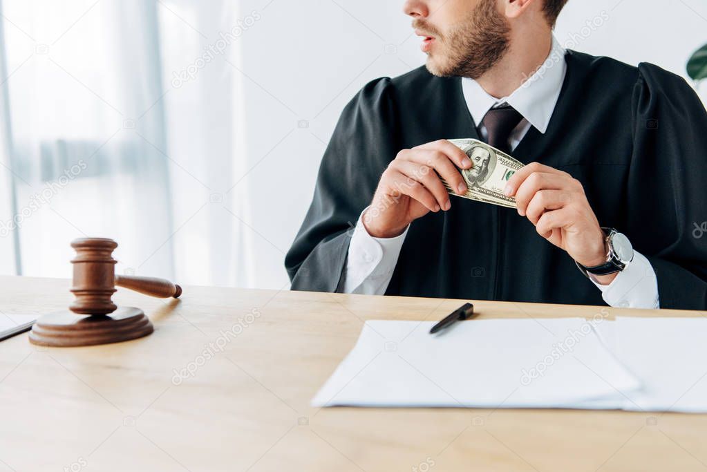 cropped view of judge holding dollar banknotes near gavel, documents and pen 