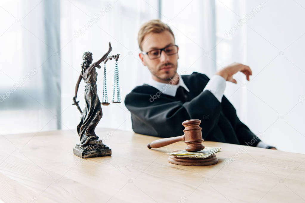 selective focus of gavel with cash and statuette of justice near judge in glasses 